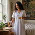 A woman stands in a softly lit bedroom wearing a white short-sleeve nightgown with lace trim and holding a glass mug. A bed, window, plants, and framed wall art are visible in the room.