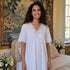 A woman with curly dark hair wearing a white short-sleeve nightgown with lace trim, standing in a bedroom in front of a tapestry and bed.