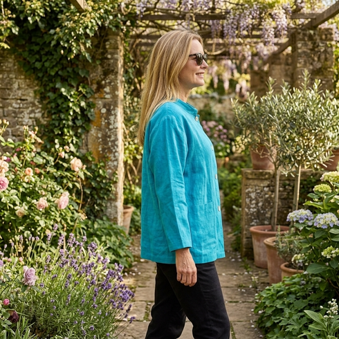 A woman wearing a bright turquoise linen jacket and black pants walks along a stone garden path surrounded by lush greenery, potted plants, and flowers.