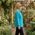 A woman wearing a bright turquoise linen jacket and black pants walks along a stone garden path surrounded by lush greenery, potted plants, and flowers.