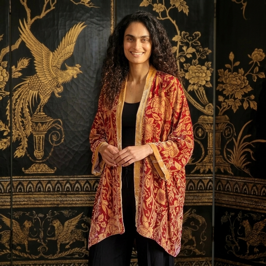A woman with long curly dark hair stands in front of a black folding screen with gold ornate patterns, wearing a red and gold patterned open-front jacket or robe over a black top and black pants.